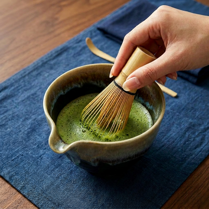 Hand whisking matcha green tea in a ceramic bowl with a wooden whisk on a blue cloth.