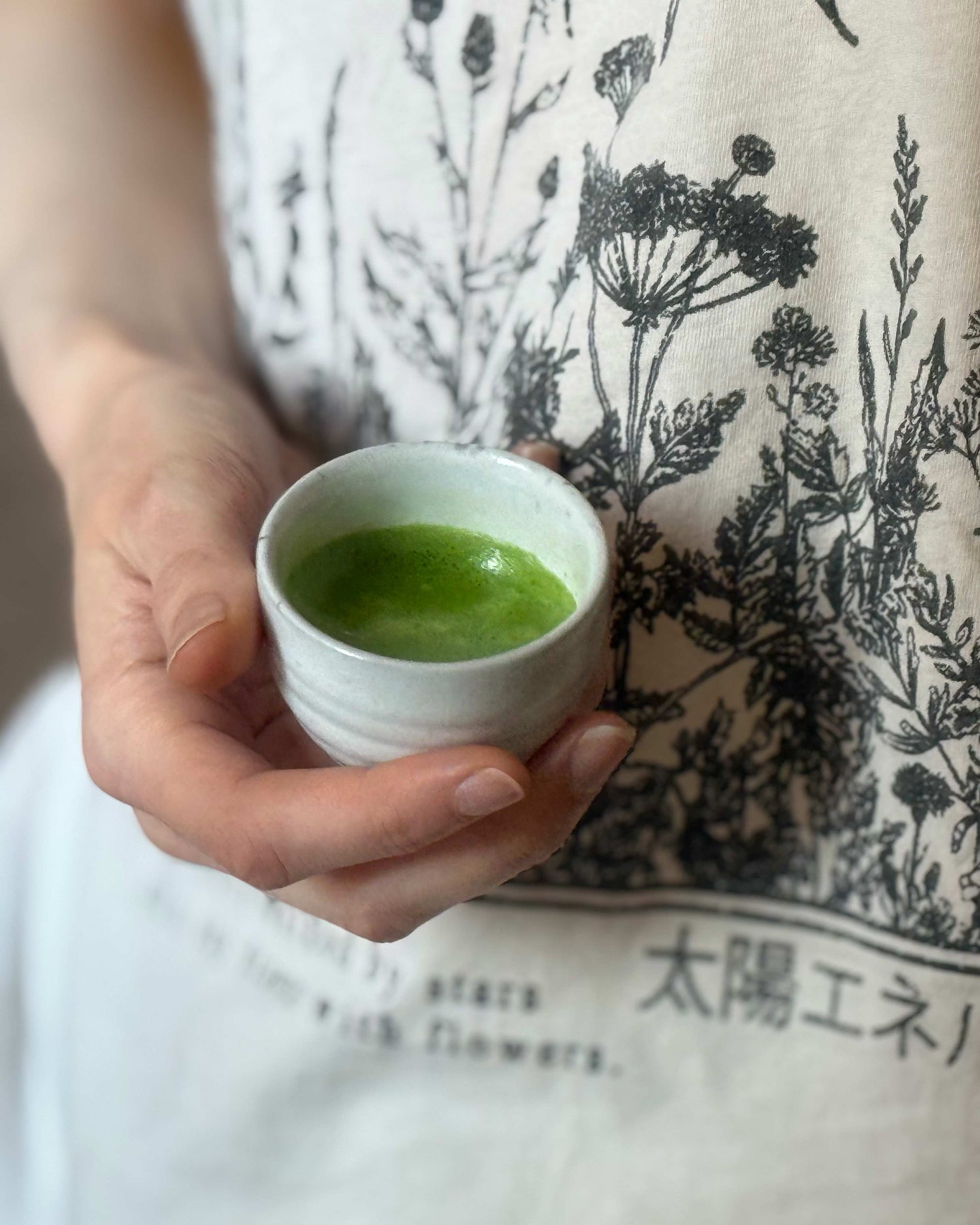 Hand holding a small white cup of matcha with a patterned shirt in the background