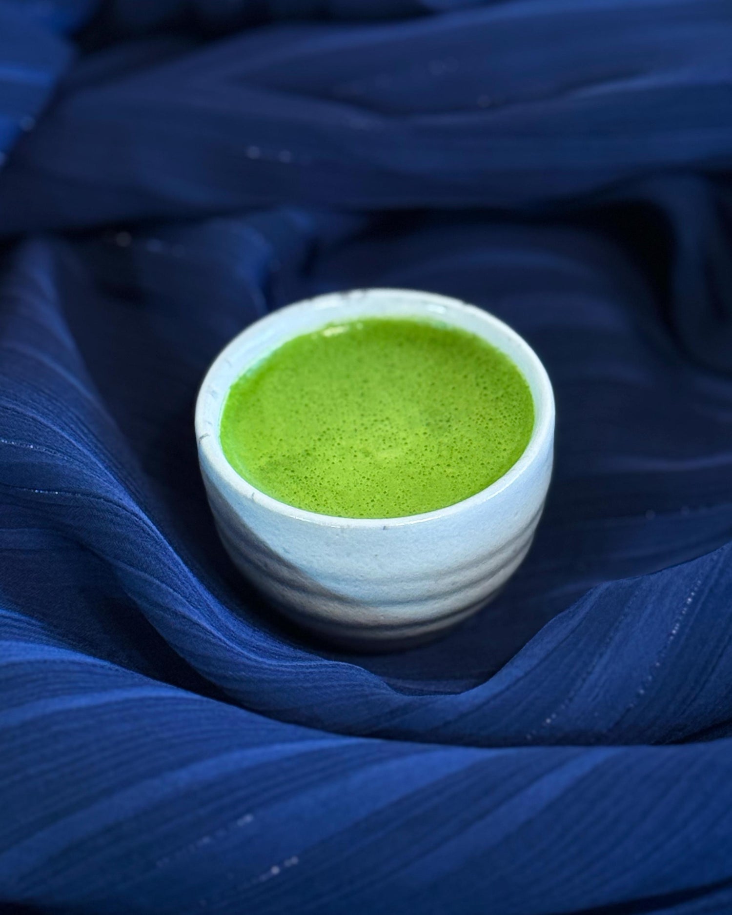 Bright green whisked matcha in a white cup on blue fabric
